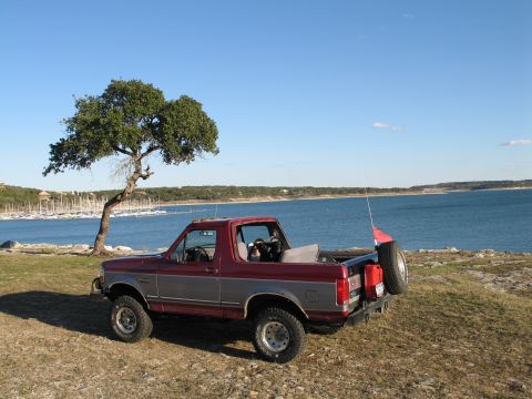 1995 Ford Bronco - A red and grey 1995 Ford Bronco is parked near a lake, with a tree and sailboats visible in the distance.