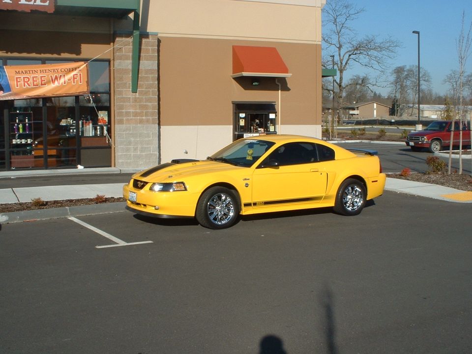 Yellow Ford Mustang parked in front of a building with a 'Free Wi-Fi' banner. Red pickup truck in background