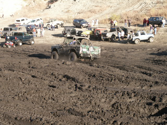 Mud-covered 1978 Ford Bronco driving through a muddy field with other vehicles and people in the background