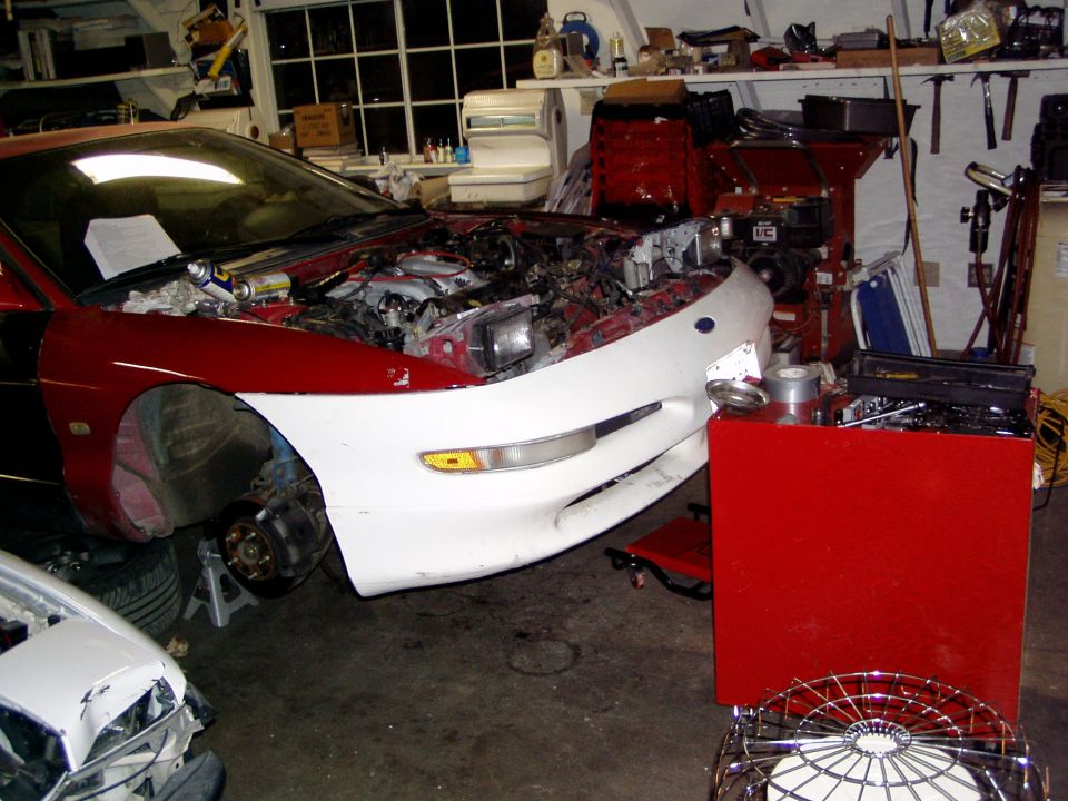 Red and white 1993 Ford Probe with hood open, front end partially disassembled, on jack stands in a garage