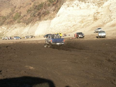1978 Ford Bronco - Multiple trucks navigate a challenging muddy terrain, with a large hill providing a backdrop to the off-road activity.