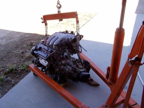 1993 Ford Probe - An engine is suspended by an orange engine hoist, with the website "SUPERMOTORS.NET" visible on the hoist's base.