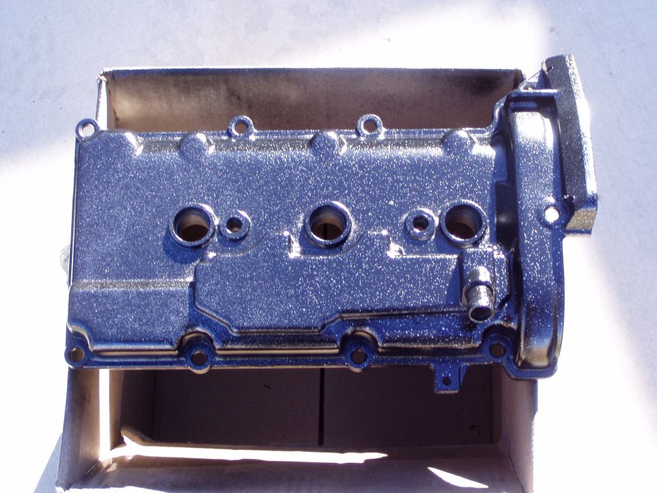 Chrome valve cover with four spark plug holes, resting in a cardboard box
