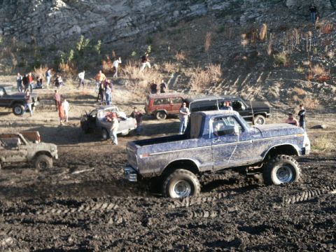 1978 Ford Bronco - A group of people and several off-road vehicles are gathered in a muddy area, with one truck stuck in the mire.