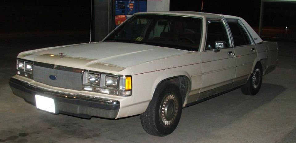 White 1980s Ford sedan, 3/4 front view, parked at a gas station