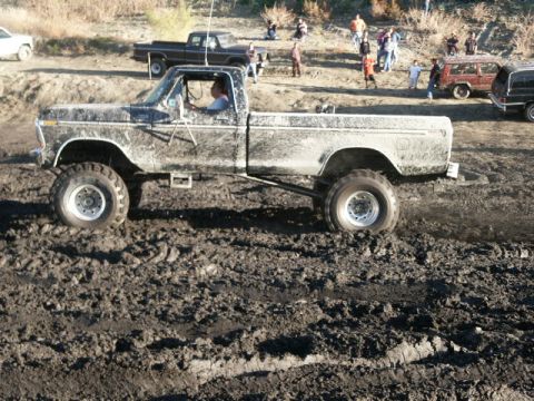 1978 Ford Bronco - A lifted pickup truck navigates a challenging muddy terrain, with spectators and other vehicles in the background.