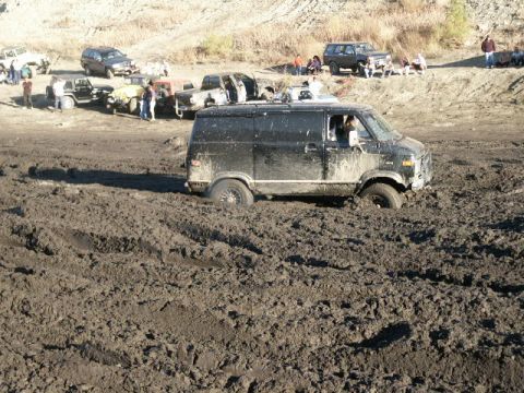 1978 Ford Bronco - A black van is stuck in thick mud, with other vehicles and people visible on higher ground in the background.