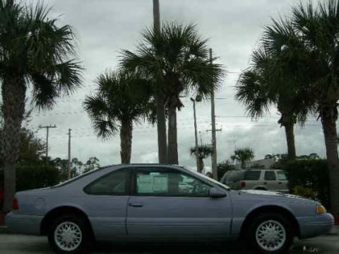 1991 Ford Bronco - A light blue coupe is parked outside with palm trees and a cloudy sky in the background.