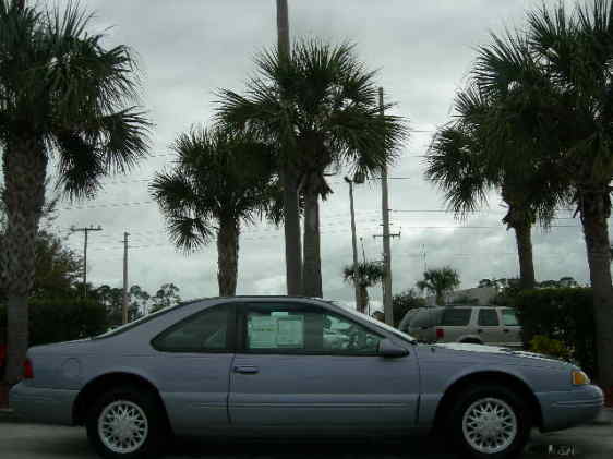 Light blue coupe with a sticker on the driver's side window, parked outdoors under palm trees