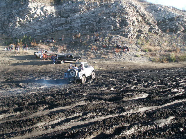 White open-top 4x4 with rear spare tire crossing deep mud ruts at an off-road area below a rocky hillside