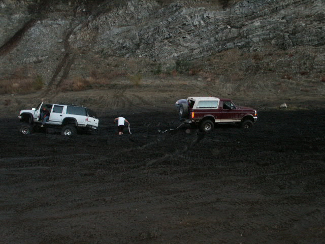 Red Ford Bronco and white lifted pickup being recovered in a black mud pit