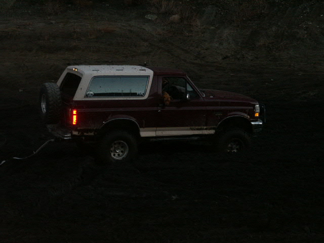 Lifted dark-red Ford Bronco with white roof and rear spare on a dirt slope at dusk