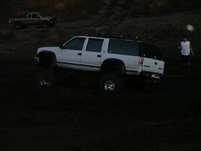 Lifted white full-size SUV with roof rack parked on dark dirt at dusk
