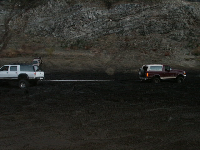 White SUV tow-strapped to a maroon Ford Bronco with white top on dark churned dirt below a rock face