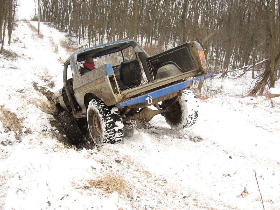 Mud-splattered blue and gray 1978 Ford Bronco struggling in snow with front wheel deep in mud