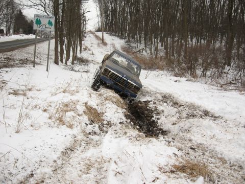 1979 Ford Bronco - A blue SUV is stuck in a muddy ditch next to a snowy road, with trees in the background.