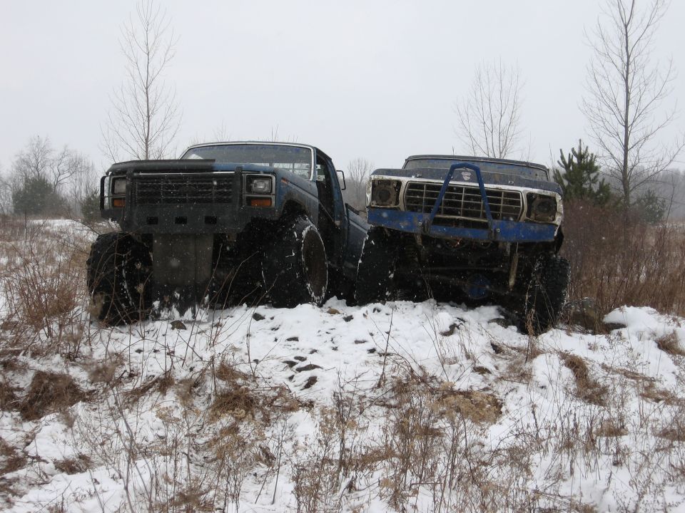 Two blue Ford Broncos with lifted suspensions and large tires in a snowy field