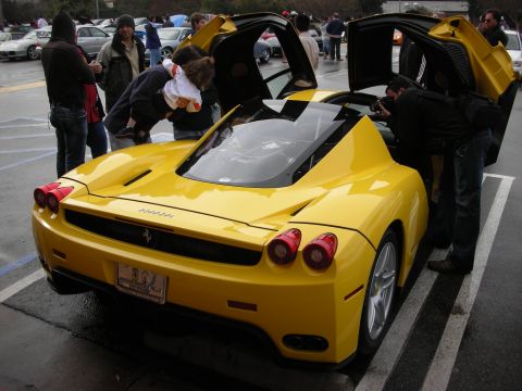 1998 Ford Crown Victoria - Yellow Ferrari Enzo seen from the rear with its scissor doors raised, surrounded by onlookers in a wet parking lot.