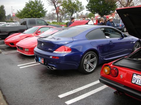 1998 Ford Crown Victoria - Blue BMW M6 coupe parked among red Ferraris on a marked lot, with people gathered in the background.