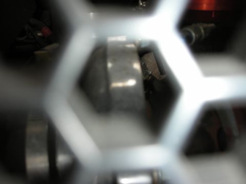 1998 Ford Crown Victoria - Engine bay hardware seen through a perforated silver hexagonal mesh panel, with a red anodized part visible at upper right.