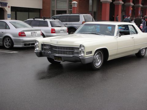 1998 Ford Crown Victoria - cream two-door 1960s Cadillac coupe with chrome grille cruises on a wet street past a shopping plaza and parked cars.