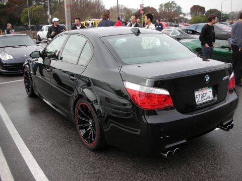 1998 Ford Crown Victoria - Rear three-quarter view of a black BMW M5 sedan with red-lipped wheels and quad exhaust in a crowded wet parking lot.