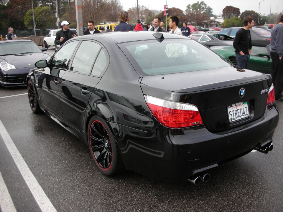 Black BMW M5 sedan rear three-quarter view in a wet parking lot with people