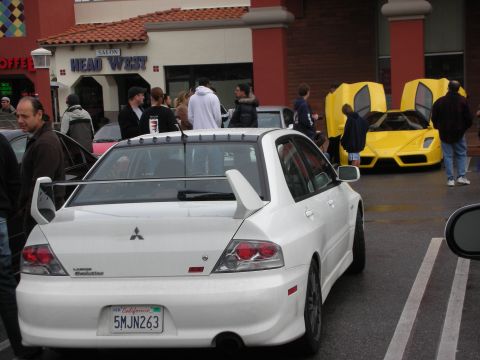 1998 Ford Crown Victoria - Rear of a white Mitsubishi Lancer Evolution with a tall rear wing in a parking lot; yellow supercars and crowd behind.