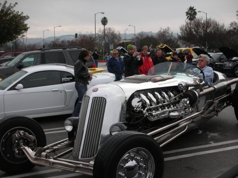 1998 Ford Crown Victoria - silver open-wheel hot rod with an exposed polished V12 engine draws a crowd in a parking lot, mountains behind.