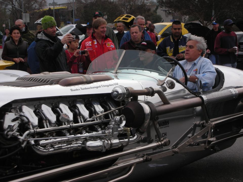 Man with white hair driving a silver V12 hot rod surrounded by spectators