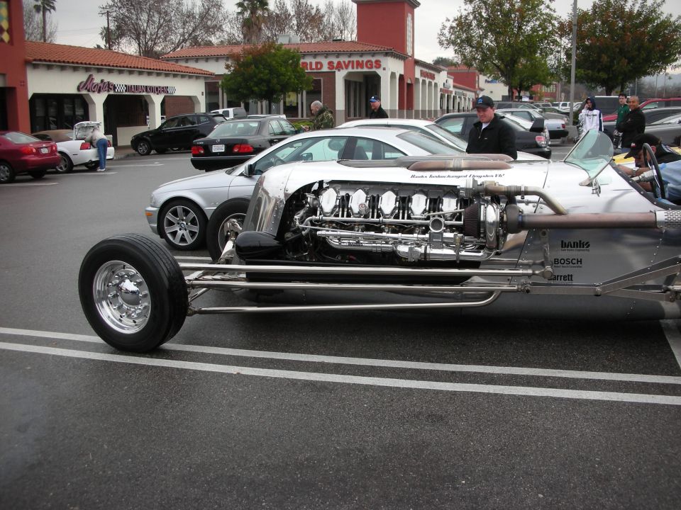 Long silver streamliner hot rod with exposed V12 engine in a parking lot