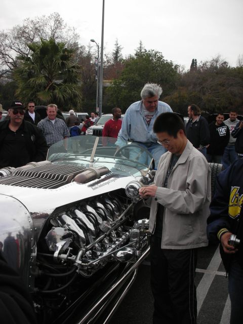 1998 Ford Crown Victoria - man in a gray jacket inspects the chrome exposed V12 of a silver hot rod while another man and a crowd look on.