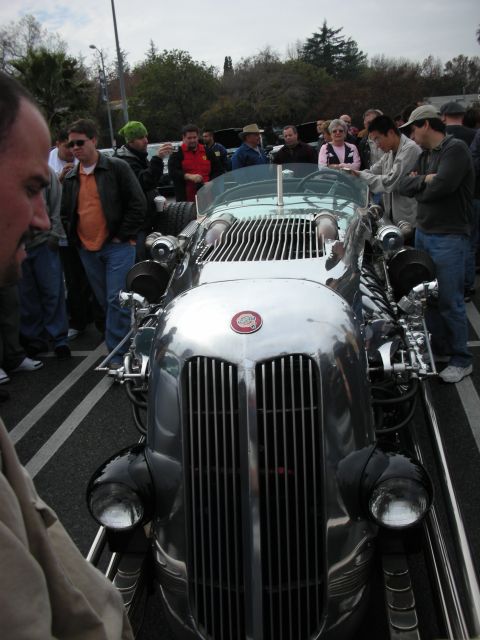 1998 Ford Crown Victoria - Head-on view of a polished silver hot rod with a tall chrome grille, round headlights, and exposed engine, crowd behind.