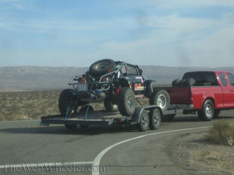 1990 Ford Bronco - black tube-chassis off-road rock buggy with a spare tire rides a flatbed trailer towed by a red pickup in the desert.