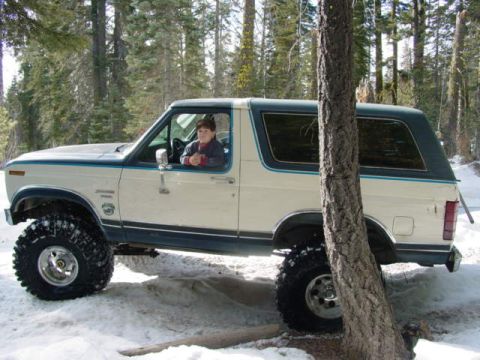 1986 Ford Bronco - A white and blue Ford Bronco with large off-road tires is parked in a snowy forest with a person in the driver's seat.