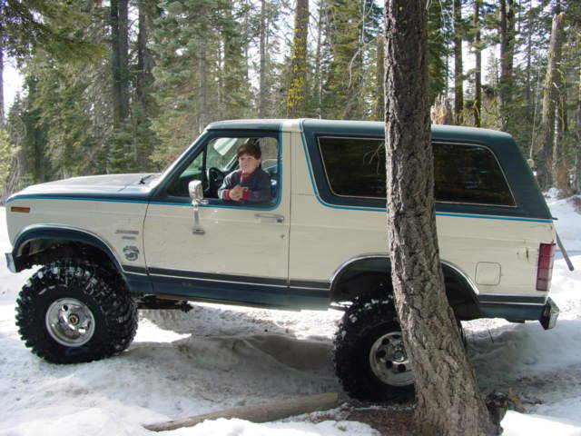 White and blue Ford Bronco with large tires in snow, driver visible