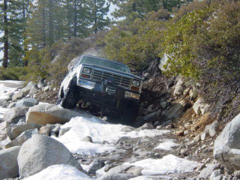 1986 Ford Bronco - A white 1986 Ford Bronco tackles a challenging off-road path with snow and rocks.