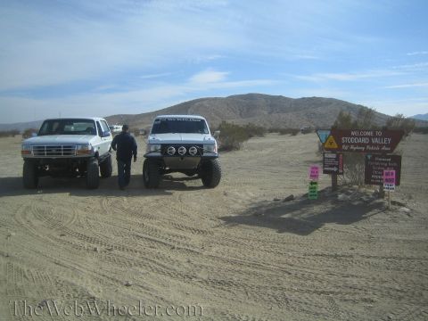 1990 Ford Bronco - Two white lifted Ford trucks, a pickup and a Bronco, parked on desert sand near the Stoddard Valley OHV sign.