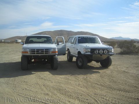 1990 Ford Bronco - white lifted Ford pickup beside a white lifted Ford Bronco with a prerunner bumper, parked on desert ground.