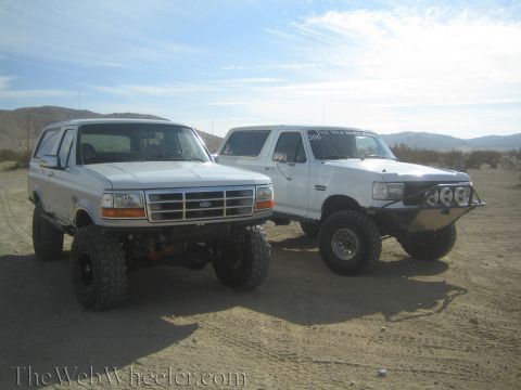 1990 Ford Bronco - white lifted Ford Bronco and a white lifted Ford pickup with prerunner lights parked side by side on desert sand.