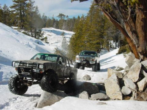 1986 Ford Bronco - Two off-road trucks navigate a snow-covered path through a wooded area.