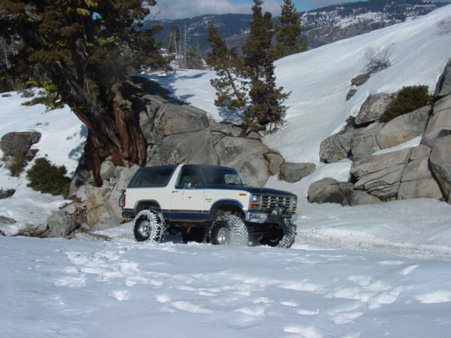 White and blue Ford Bronco with large tires driving in snow