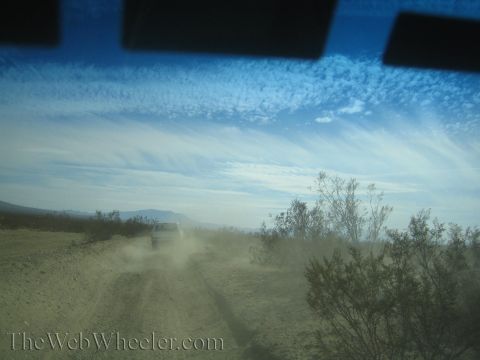 1990 Ford Bronco - white vehicle kicks up dust on a desert trail beneath a wide sky of streaky clouds, seen through a windshield.