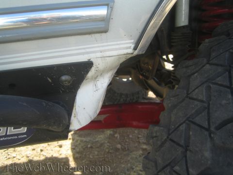 1990 Ford Bronco - Detail of a white off-road SUV's rocker panel and red lower control arm above a mud-terrain tire on rocky desert ground.