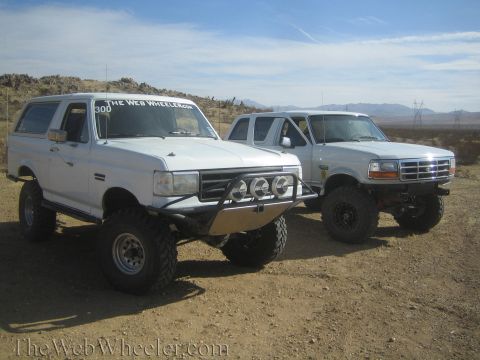 1990 Ford Bronco - white lifted Ford Bronco with light bar bumper parked beside a silver lifted Ford Bronco in the desert.