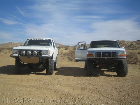1990 Ford Bronco - Front view of a white and a silver lifted Ford Bronco parked side by side in the open desert under a blue sky.