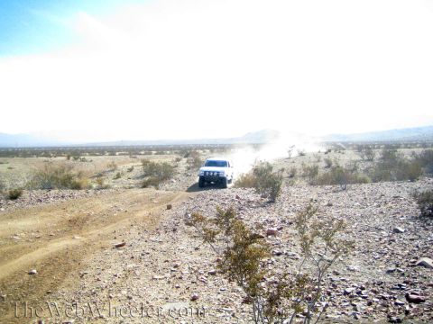 1990 Ford Bronco - white off-road vehicle drives along a rocky desert trail, trailing a cloud of dust under a hazy sky.