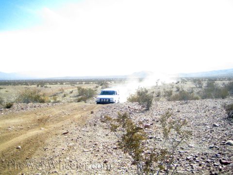 1990 Ford Bronco - white off-road vehicle with a light bar drives a dusty rocky desert track toward the camera.