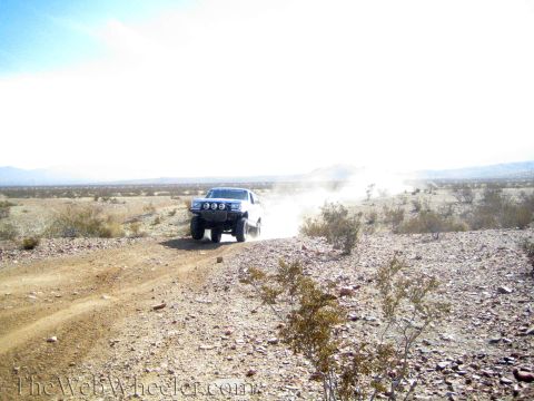 1990 Ford Bronco - white lifted truck with auxiliary lights kicks up dust driving toward the camera on a desert dirt trail.
