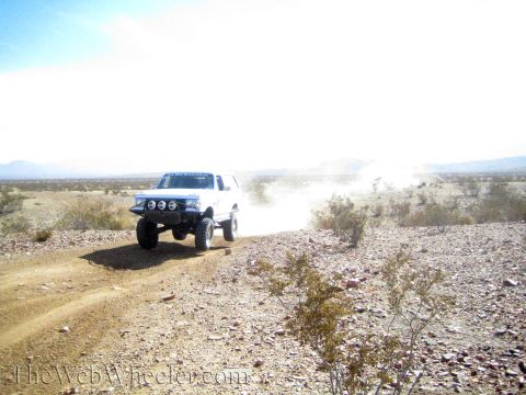 1990 Ford Bronco - white lifted SUV with off-road lights crests a dusty desert ridge, trailing dust across rocky terrain.